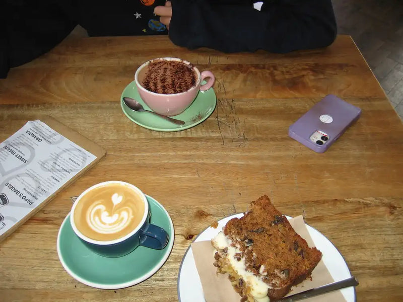 Two cups of coffee and a slice of cake on a wooden table with a phone and newspaper at swim club afters