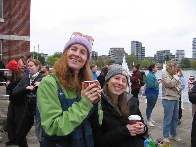 Two women holding hot drinks by London's West Reservoir with people and buildings in the background after a winter open water swim