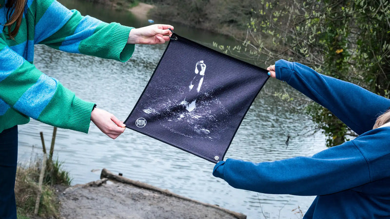 Two people holding a Swim Peeps Dry Step Swim Changing Mat outdoors by an outdoor swimming pond