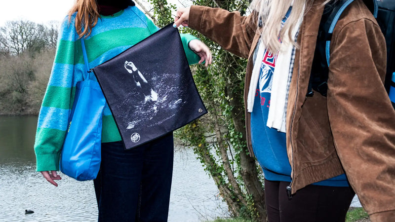 Two people standing outdoors by a body of water, one holding up the corner of a Swim Peeps Dry Step Swim Changing Mat