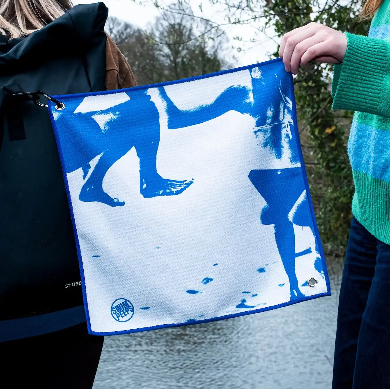 Two people standing by a swimming pond, one holding up the corner of a Dry Step Swim Changing Mat