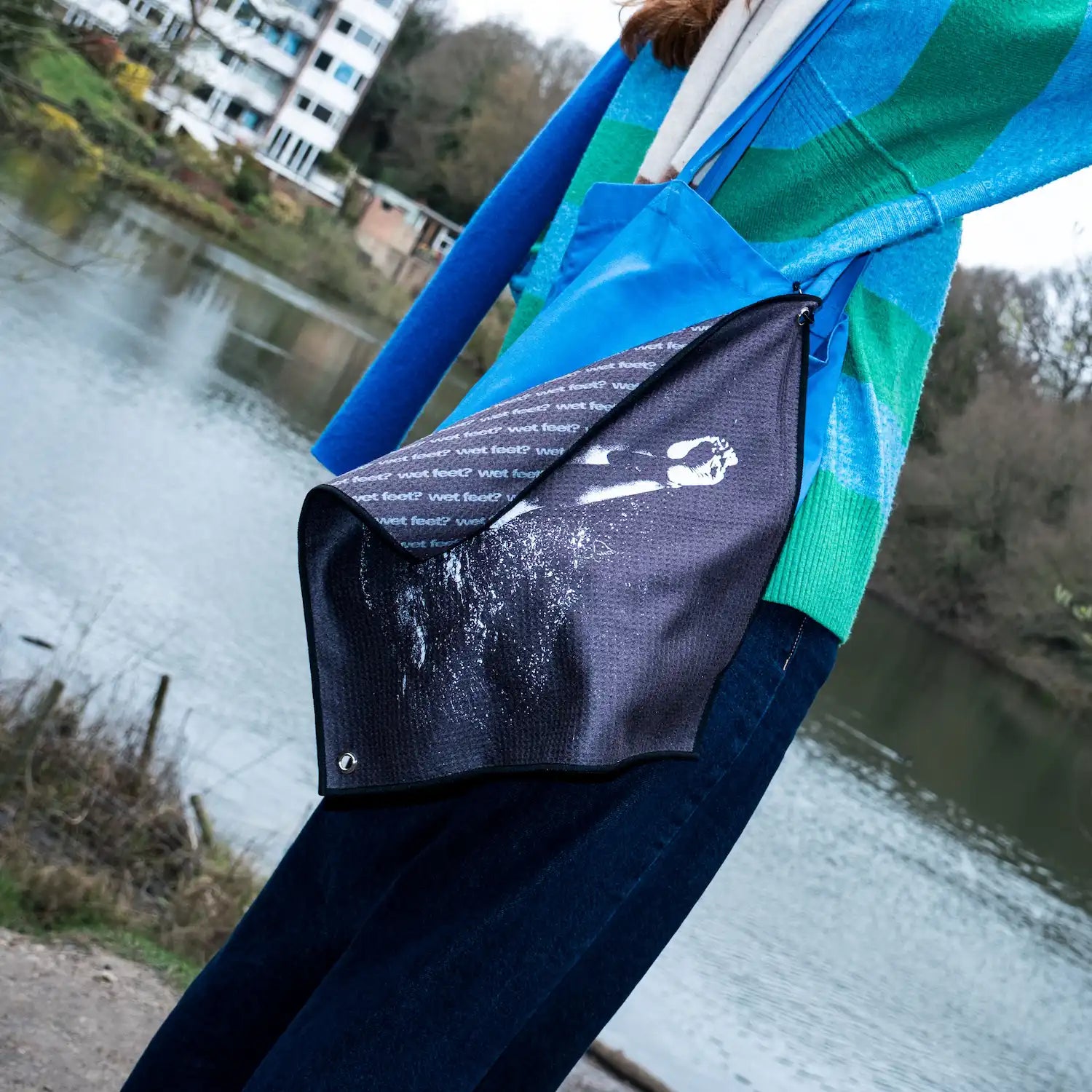 The Swim Peeps Dry Step Swim Changing Mat hanging from a tote bag in front of a swimming pond